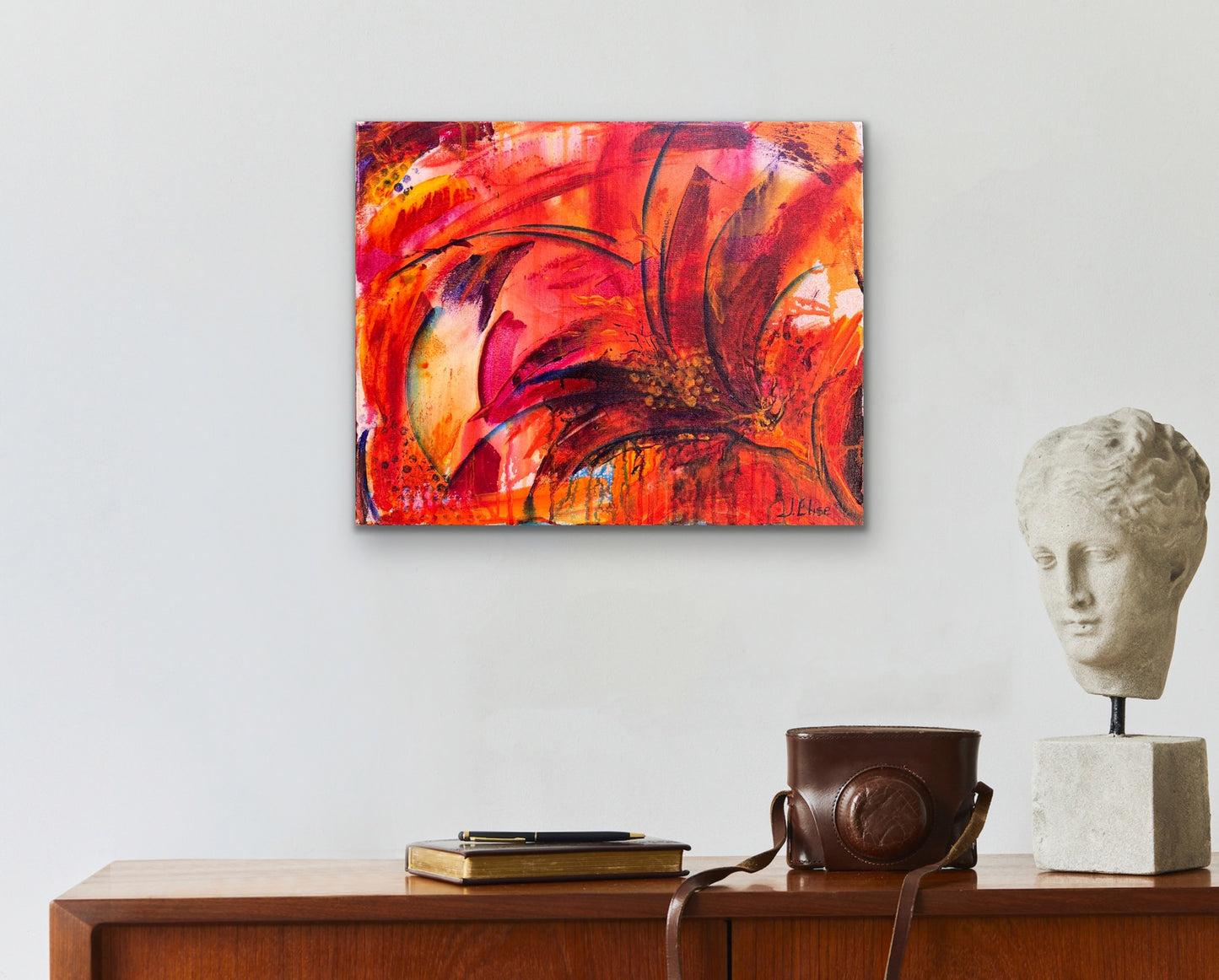 In situ photo of fiery abstract floral artwork with bold red, orange, and magenta tones, featuring dynamic petal shapes and expressive brushwork that radiates movement and vitality in an academia style home interior on a white wall.