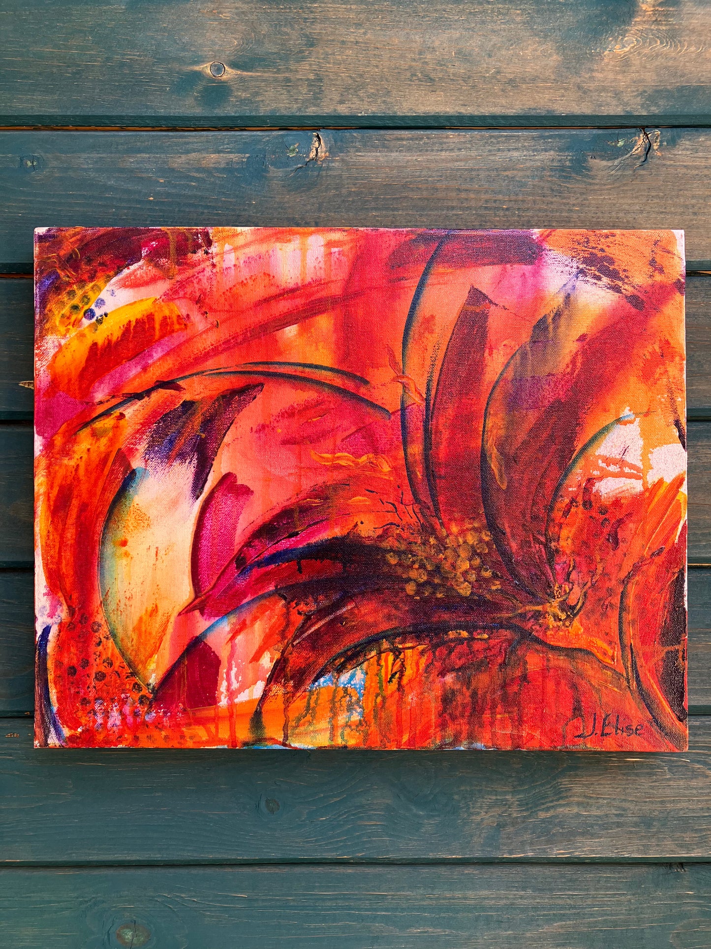 Fiery abstract floral artwork with bold red, orange, and magenta tones, featuring dynamic petal shapes and expressive brushwork that radiates movement and vitality on a rustic wood siding wall.