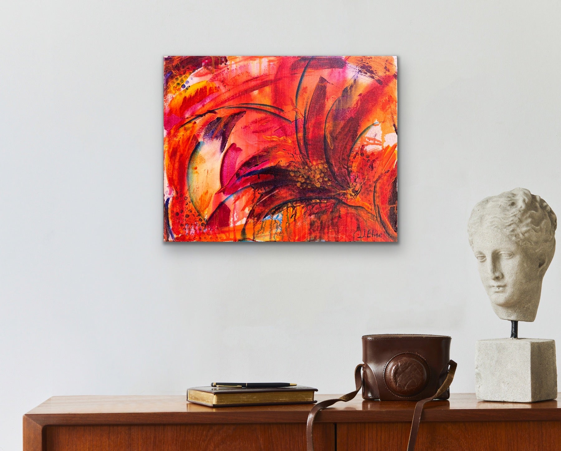 In situ photo of fiery abstract floral artwork with bold red, orange, and magenta tones, featuring dynamic petal shapes and expressive brushwork that radiates movement and vitality in an academia style home interior on a white wall.