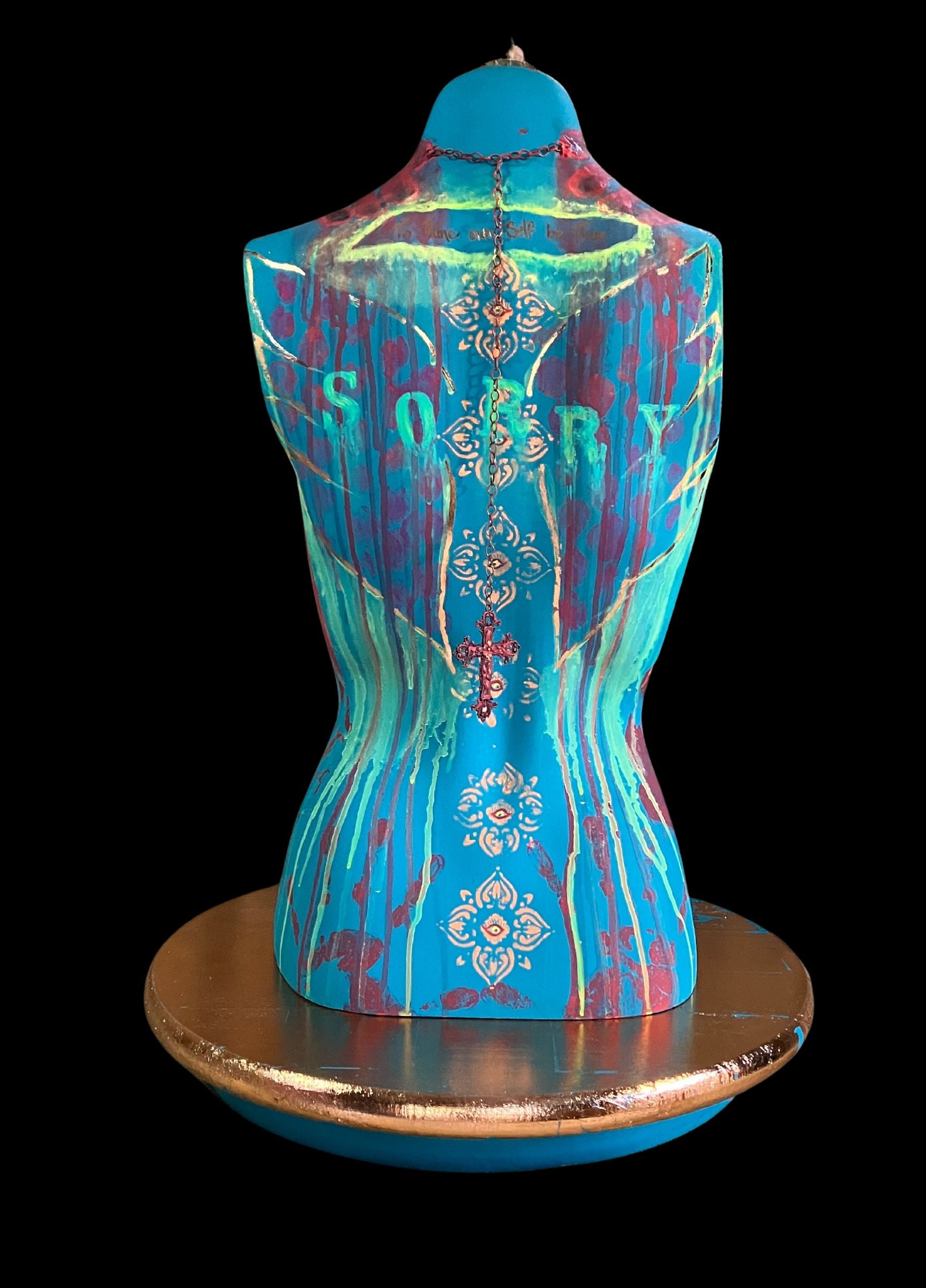 The back of a mixed media mannequin sculpture titled "Break Free", featuring bold turquoise body, rose gold guilded wings, the word SORRY appearing to melt away, the phrase "To thine own self be true" in rose gold, a cross hanging from a long single chain hanging from a choker necklace and a turning base by J Elise Art.