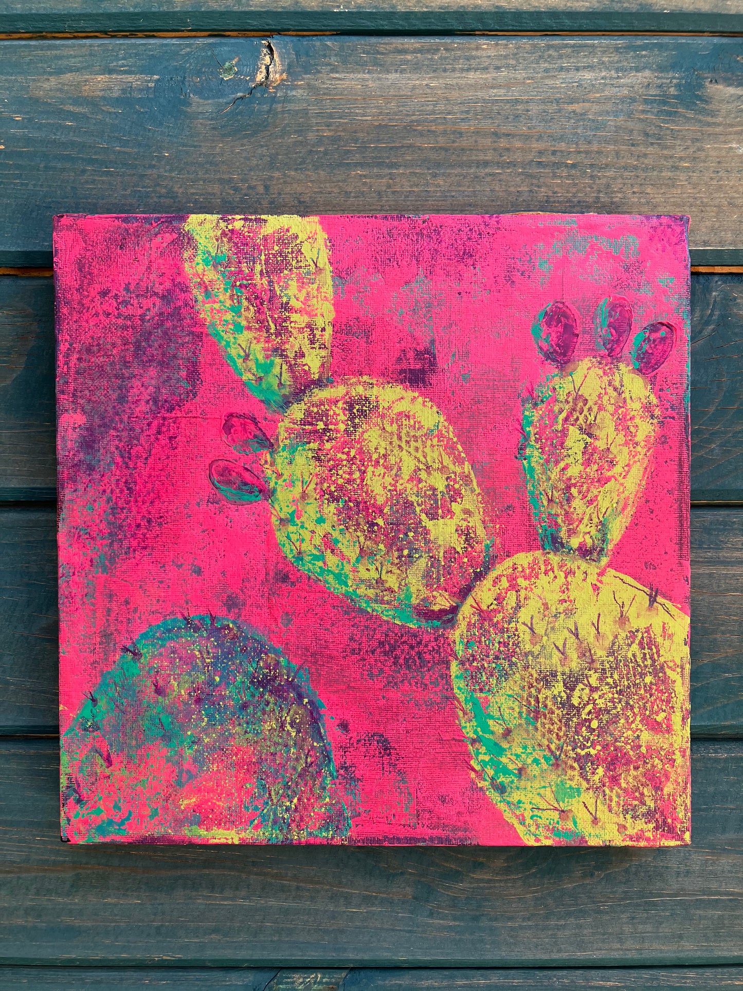 Bright abstract desert cactus painting with neon yellow prickly pear pads on a vivid pink background, symbolizing new growth and early spring bloom o a rustic wood slat wall.